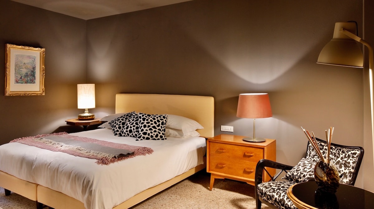 A comfortably arranged bedroom features a spacious bed with a patterned throw and soft pillows. Ambient lighting is provided by a bedside lamp and a stylish floor lamp. A wooden nightstand and a chair with a unique design add to the room's character.