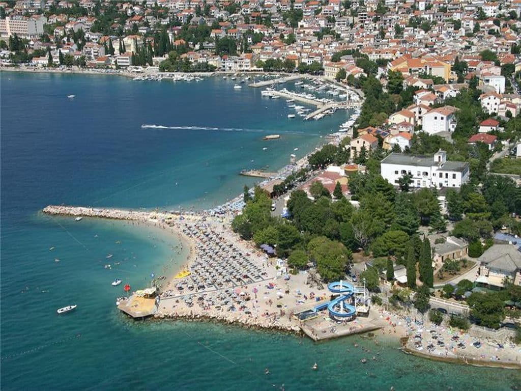 An aerial view captures the coastal town, highlighting the sandy beach filled with sunbathers and umbrellas. Clear blue waters surround the area, along with a marina displaying anchored boats. Lush greenery lines the beach, offering a natural contrast to the urban landscape in the background.