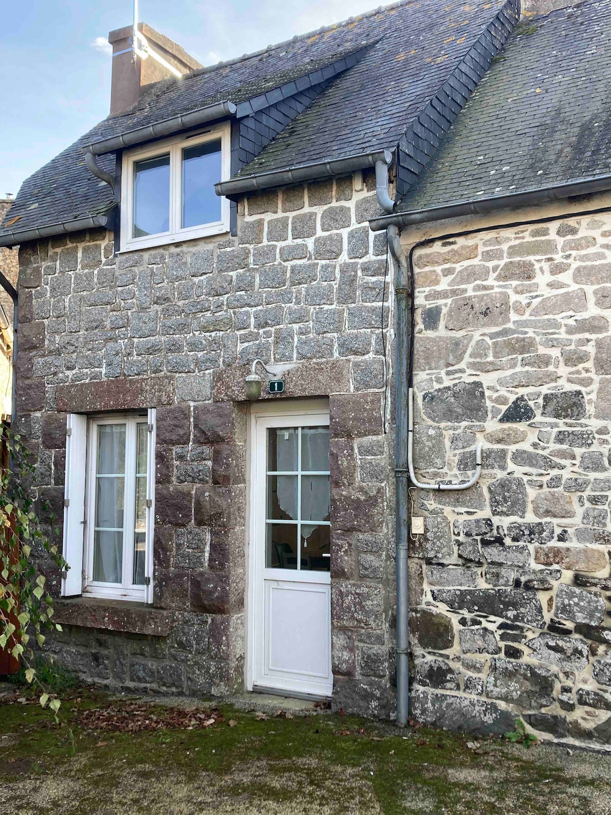 A charming stone cottage exterior is showcased, featuring a combination of light and dark stones. Two windows are positioned symmetrically on the upper level, while a white door leads to the entrance. Nearby, creeping greenery adds a touch of nature to the front.