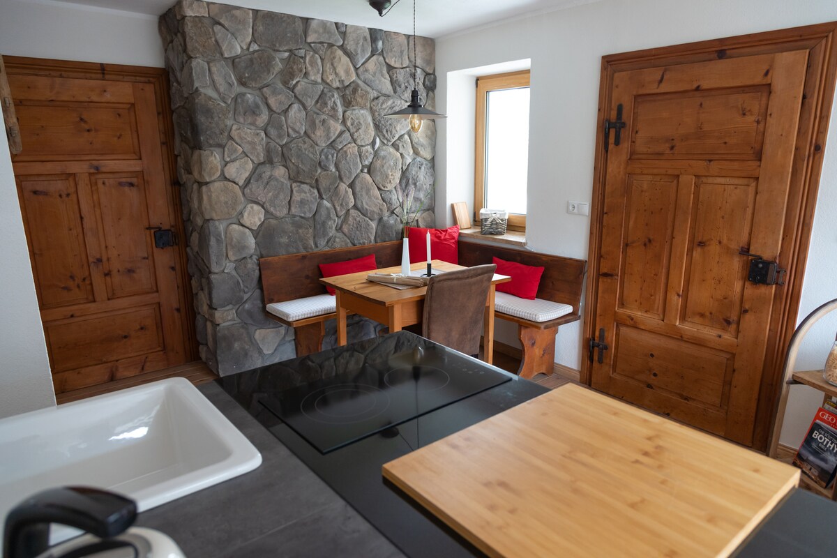 A cozy kitchen area features a natural stone wall and wooden furniture. A small wooden table is surrounded by a comfortable corner bench and additional chairs. Sunlight enters through a window, illuminating the warm wood tones of the walls and doors.