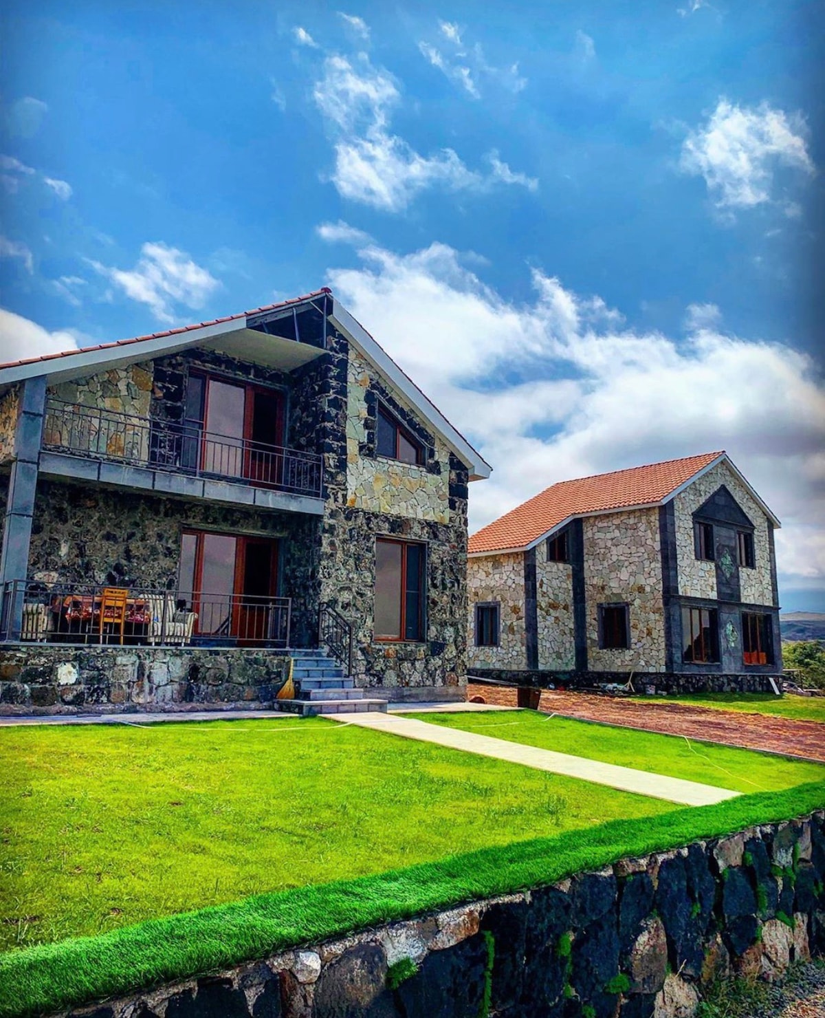 Two stone houses are set in a lush green landscape, with expansive windows reflecting the sky. A pathway leads from the foreground to the homes, framed by a well-manicured lawn. The clouds softly enhance the backdrop of this serene setting.