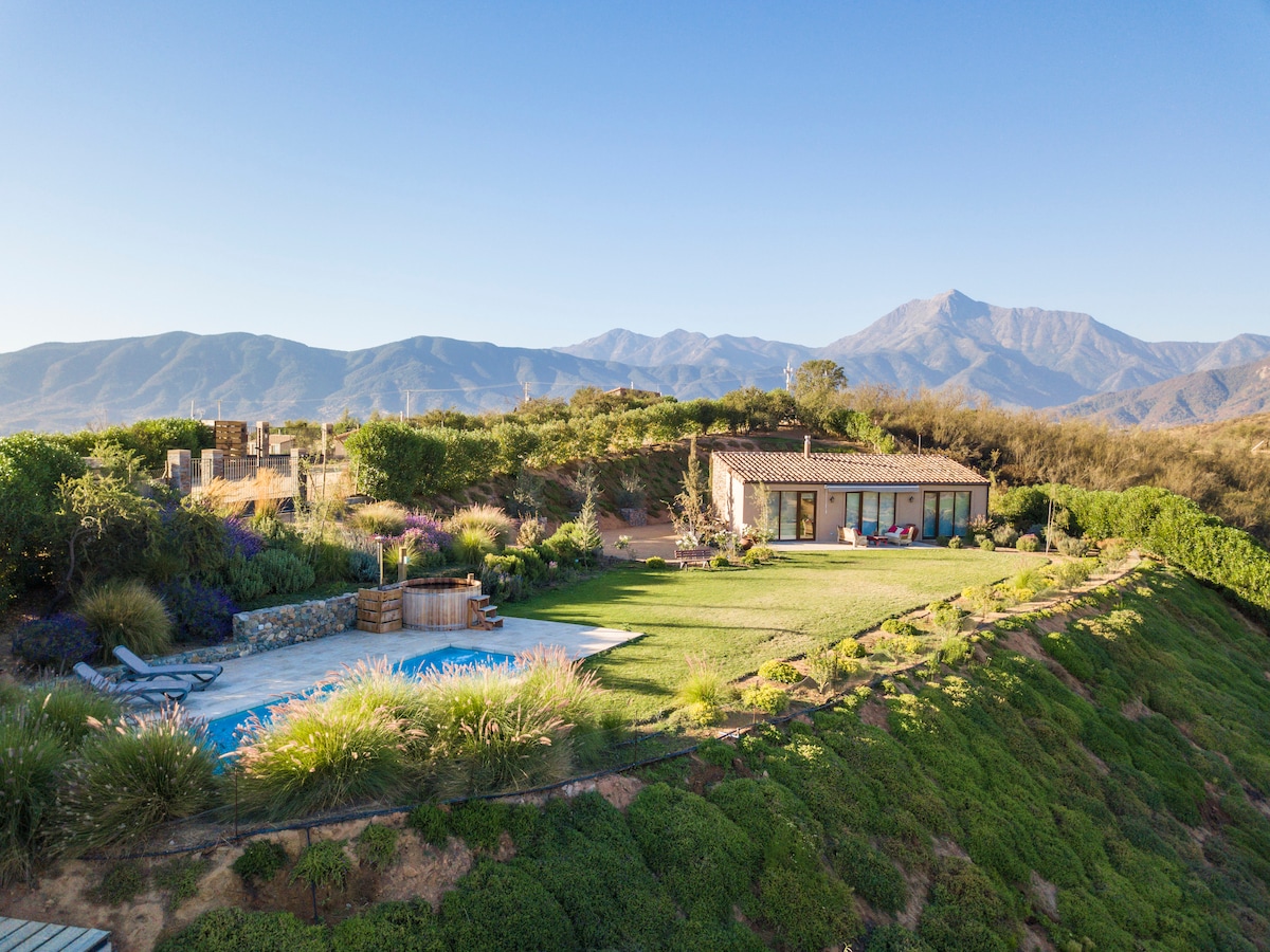 A spacious villa is set against a backdrop of mountains, surrounded by lush green landscaping. Inviting areas for relaxation are provided, including a swimming pool and a hot tub, while soft hills and flora enhance the tranquil outdoor environment.