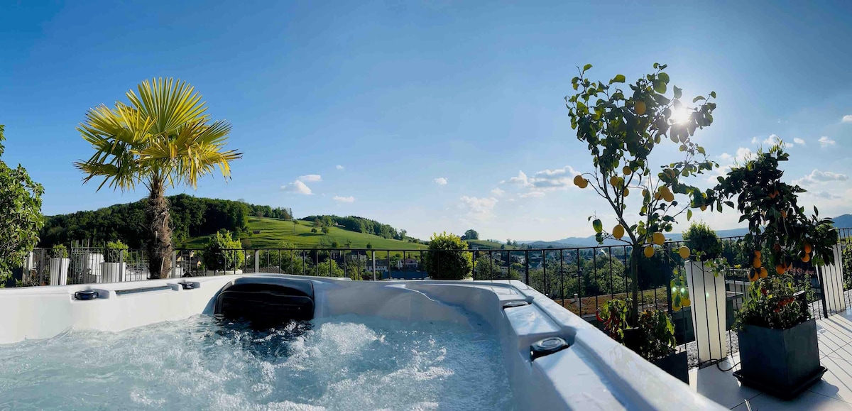 A hot tub with bubbling water is positioned on a terrace surrounded by various potted plants, including a palm tree and citrus trees. The expansive landscape is visible in the background, showcasing lush green hills under a clear blue sky with soft sunlight.