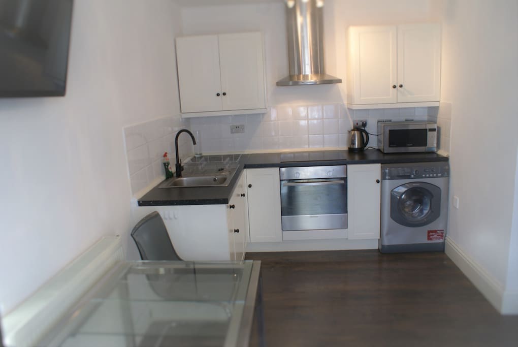 Kitchen with oven, washing machine, microwave, plates, glasses, mugs and cutlery, sink and hob. Wall mounted TV on right