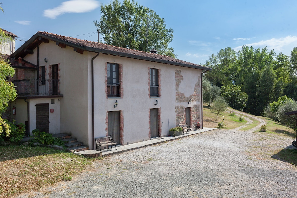 A two-story farmhouse is set against a tranquil landscape. The exterior features a mix of rustic stone and plastered walls, complemented by wooden shutters. A gravel parking area is visible in the foreground, leading to a tree-lined path that extends into the greenery.