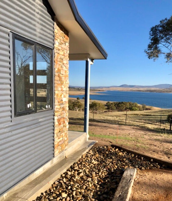 The exterior of the chalet is showcased, featuring a stone finish and large windows. The serene view of Lake Jindabyne is visible in the background, framed by gentle hills under a clear blue sky. A pebbled area leads to the entrance.
