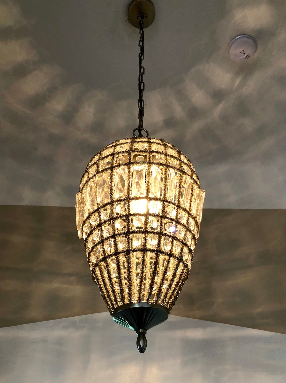 A stunning chandelier hangs from the ceiling, featuring a detailed design with shimmering crystals. Light reflects softly from its surface, casting intricate patterns on the surrounding ceiling, enhancing the room's elegance.