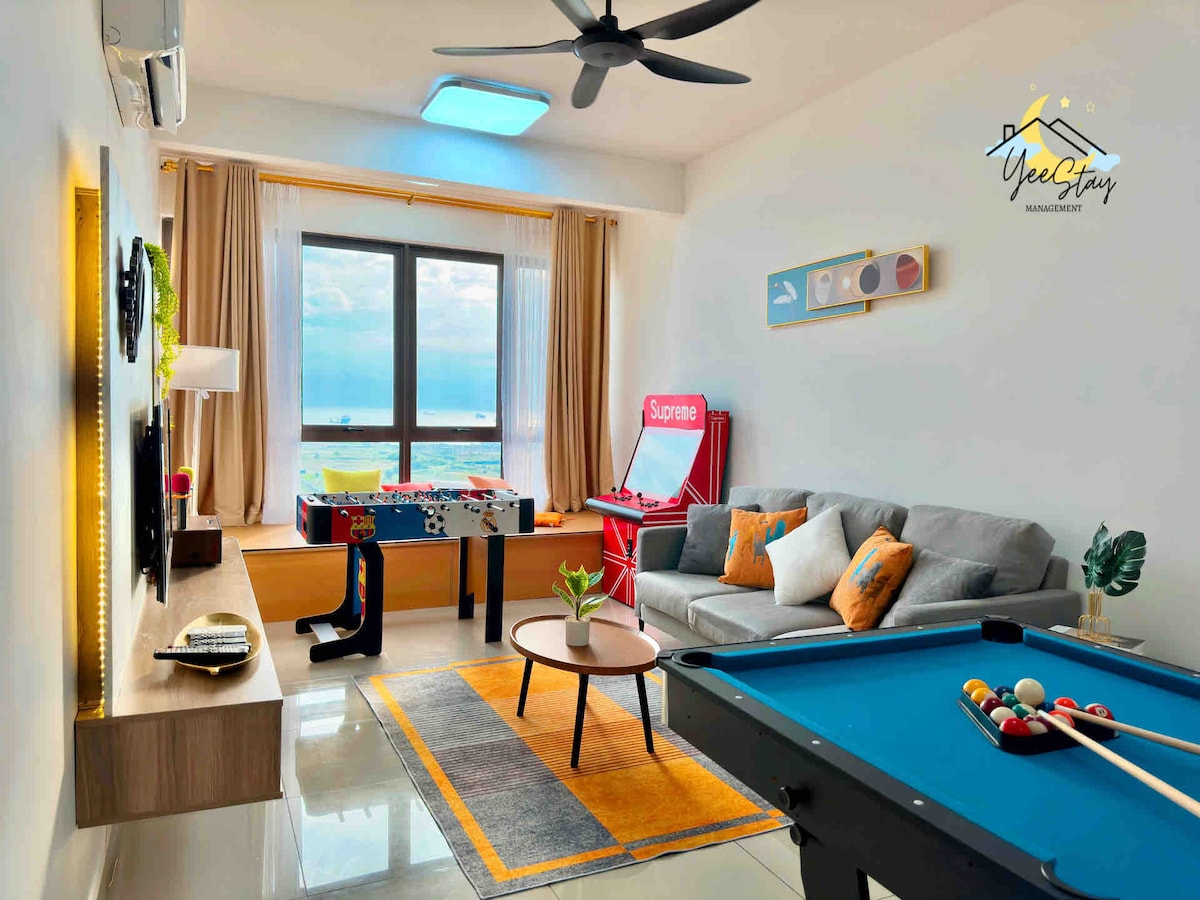 A spacious living room showcases a comfortable sofa set, a coffee table, and a vibrant area rug. A pool table and an arcade game machine add entertainment options. Large windows allow natural light and present a view of the sea, while soft curtains frame the glass.