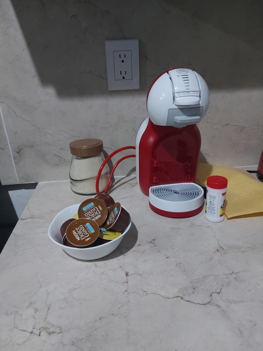 A red coffee machine sits on a light-colored countertop, accompanied by a glass jar of sugar and a bowl containing coffee pods. A yellow kitchen towel and a small bottle of seasoning are also visible, adding practical elements to the space.