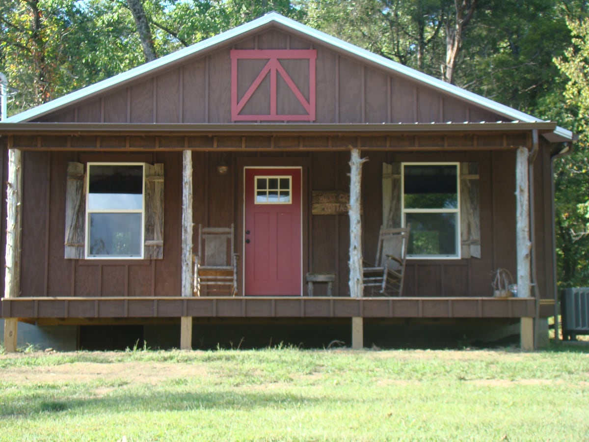 The cottage exterior features a welcoming covered front porch with two wooden rocking chairs. A red door adds a pop of color, complemented by the brown barn wood walls. Two large windows on either side of the door allow natural light to enter, enhancing the cottage's charm.