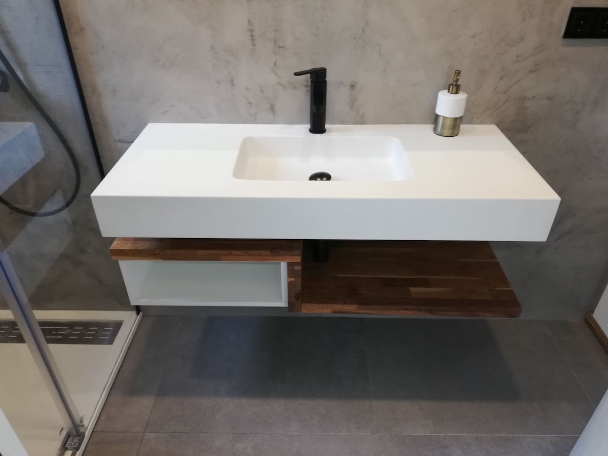 A modern bathroom sink area is presented, featuring a sleek white countertop with an integrated basin. Below, a wooden shelf is positioned on one side, while a small storage compartment is visible on the other. The wall is finished in a muted gray texture, enhancing the contemporary design.