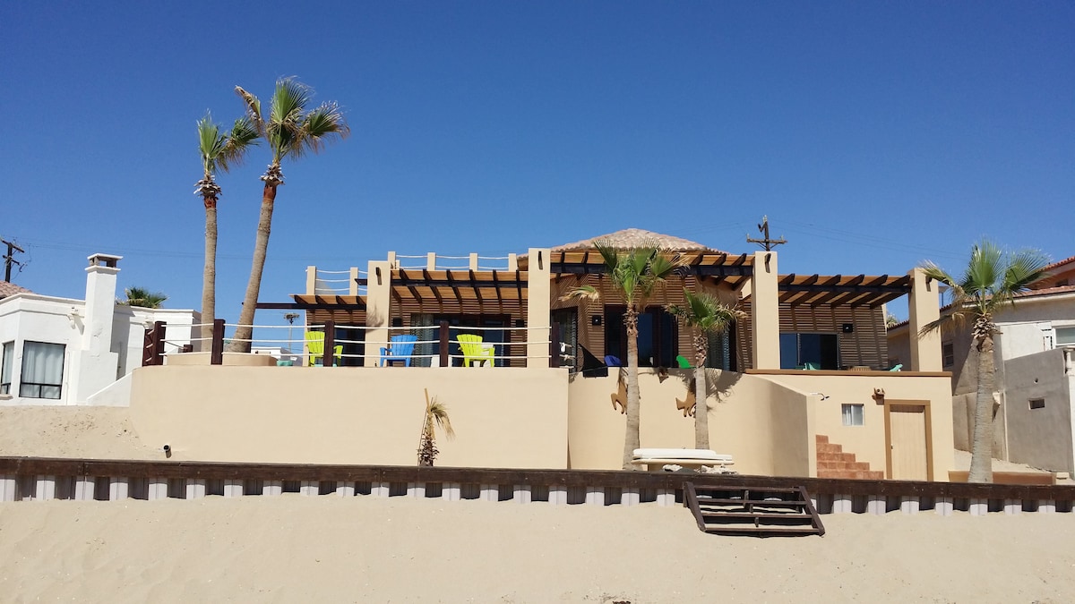 A beachfront home is presented with a modern architectural style, featuring a terracotta tiled roof and a spacious covered patio. Palm trees surround the property, and colorful chairs are arranged outside for enjoying the view. The sandy beach leads up to the entrance, enhancing the coastal ambiance.