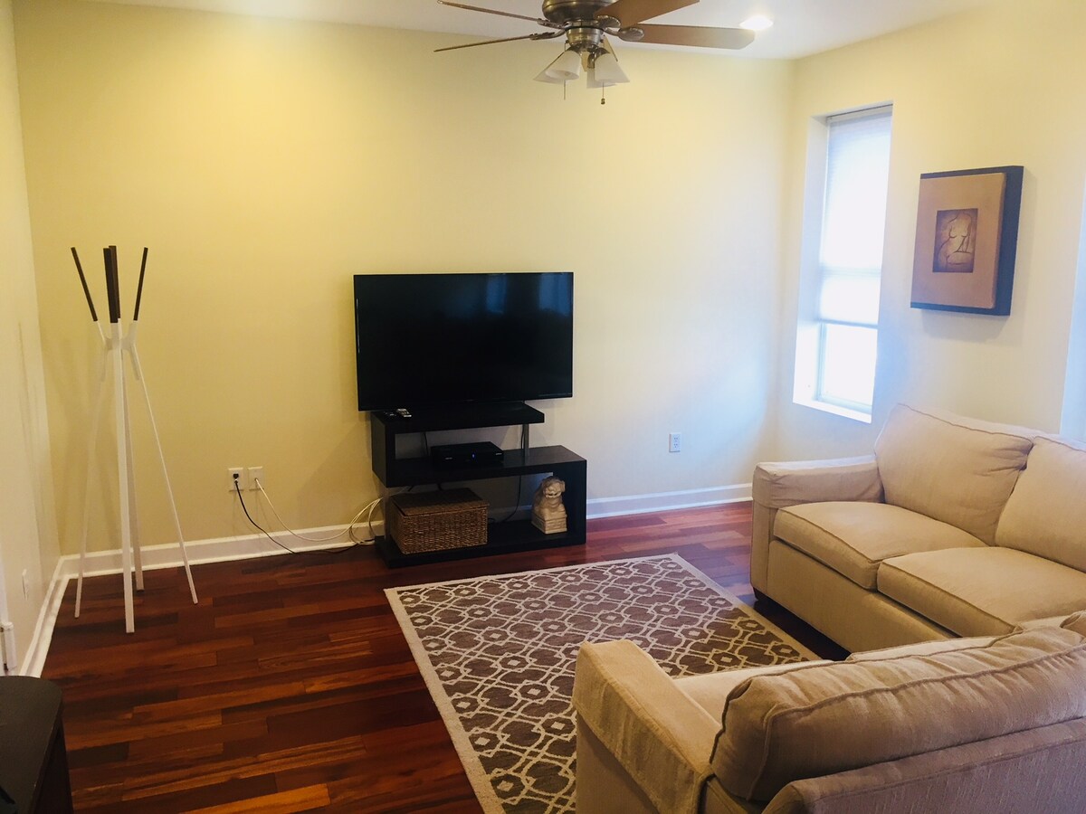 A living area is featured, with a light yellow wall providing a soft backdrop. A cream-colored couch is positioned opposite a black media console holding a flat-screen television. A patterned rug lies on hardwood flooring, accompanied by a single window, allowing natural light to fill the space.