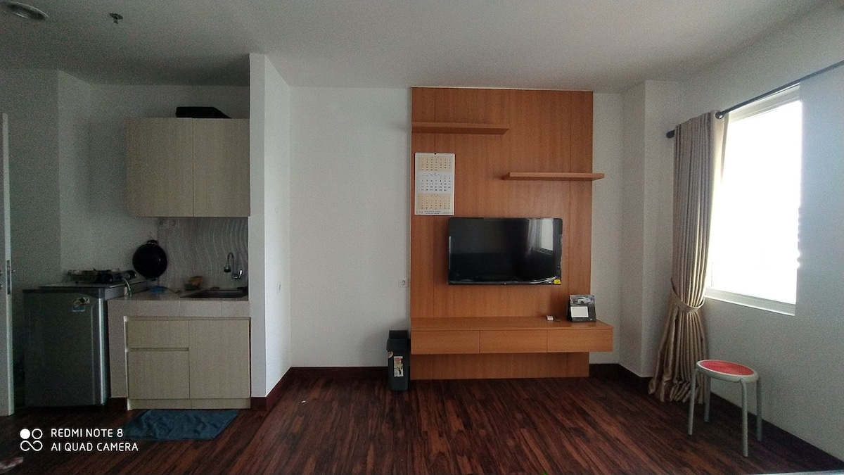 A modern living space is featured, showcasing a wooden wall-mounted TV unit. Kitchen amenities are present in a light-colored cabinet adjacent to the living area. A small red chair provides a pop of color, while natural light filters through the large window, framed by beige curtains.
