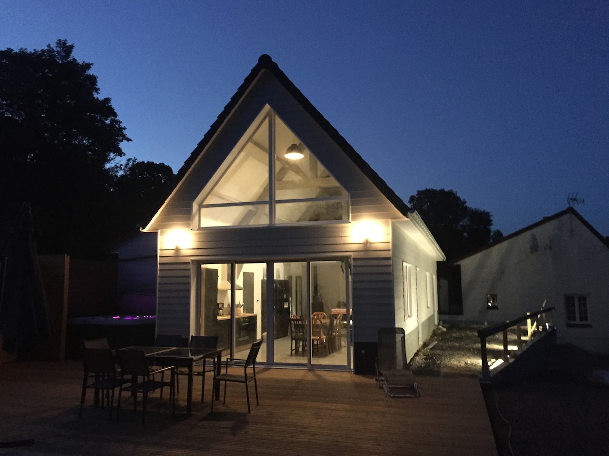 The exterior of a modern gîte is shown in the evening light, featuring a gabled roof and large glass doors. Soft illumination highlights the architecture, while outdoor seating is arranged on a spacious deck. A glimpse of the spa area is visible in the background.