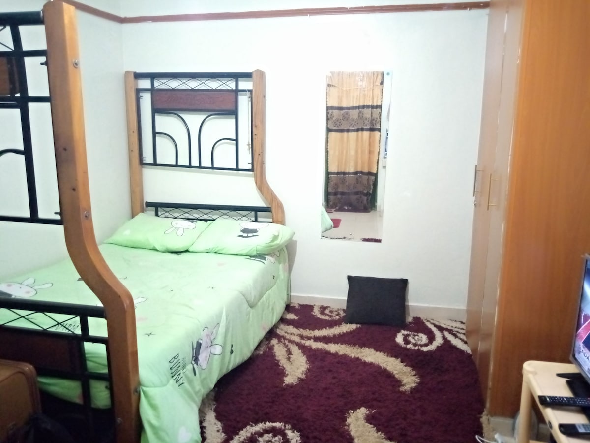 A cozy room features a bunk bed with a green bedding set. A mirror reflects the interior, while a dark pillow is placed on a decorative rug. A television is visible on a stand opposite the bed, with a wardrobe providing additional storage.