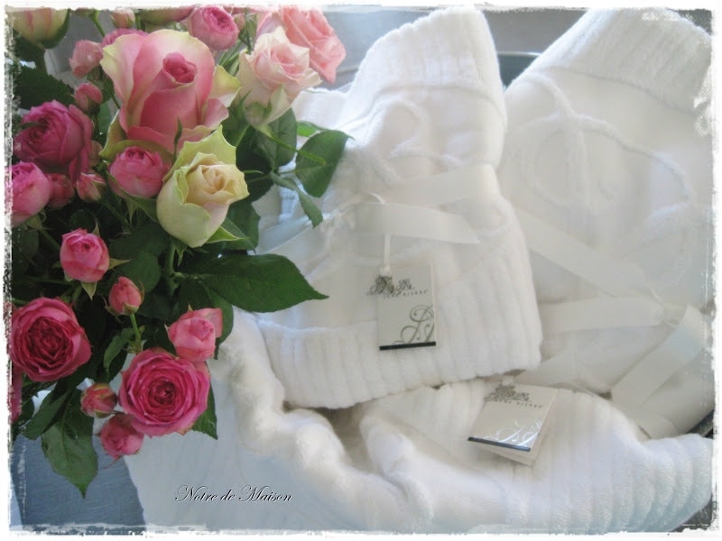 Soft white bathrobes are displayed alongside a bouquet of pink and cream roses. The robes are neatly folded with tags attached, while the arrangement of flowers adds a touch of elegance to the setting.