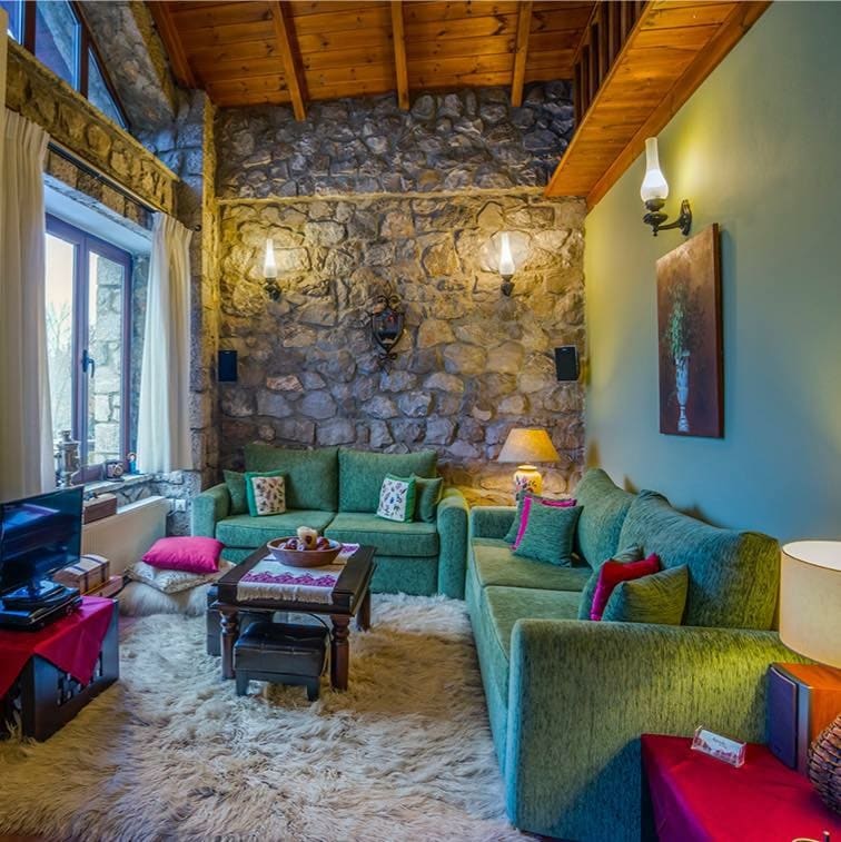 A stone wall enhances the rustic charm of this living area, where two green sofas and a wooden coffee table create a cozy seating arrangement. Soft rugs cover the floor, while natural light enters through large windows, complemented by warm lighting fixtures.
