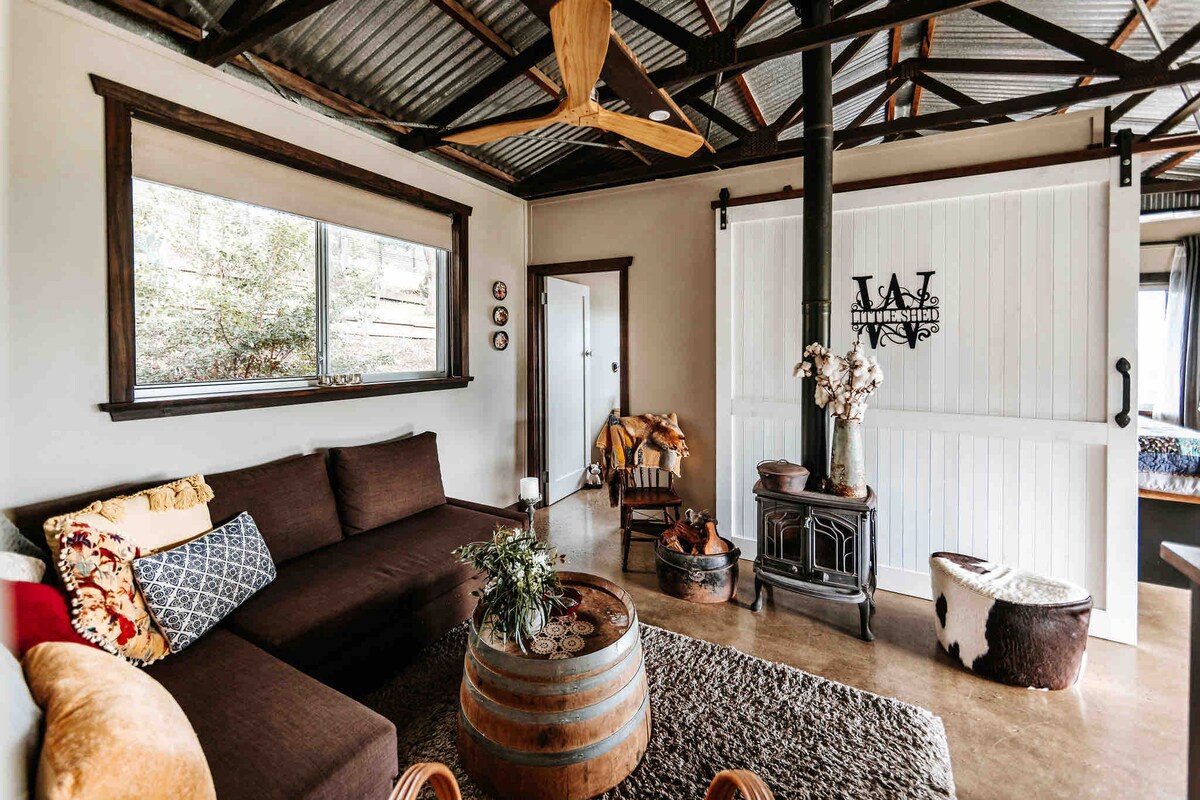 The living area features a dark brown sofa against a light-colored wall, complemented by a wooden coffee table made from a barrel. A cast iron fireplace stands nearby, and a ceiling fan is mounted above. Natural light enters through a large window overlooking the surrounding greenery.