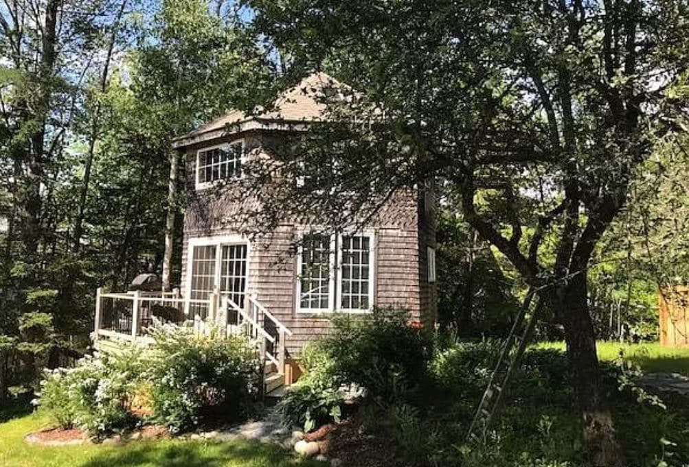 A hexagonal cottage is nestled among trees in a lush green landscape. Windows are framed with natural wood, allowing for ample light. A wooden deck leads to the entrance, surrounded by vibrant plant life. The setting offers a serene and welcoming outdoor space.