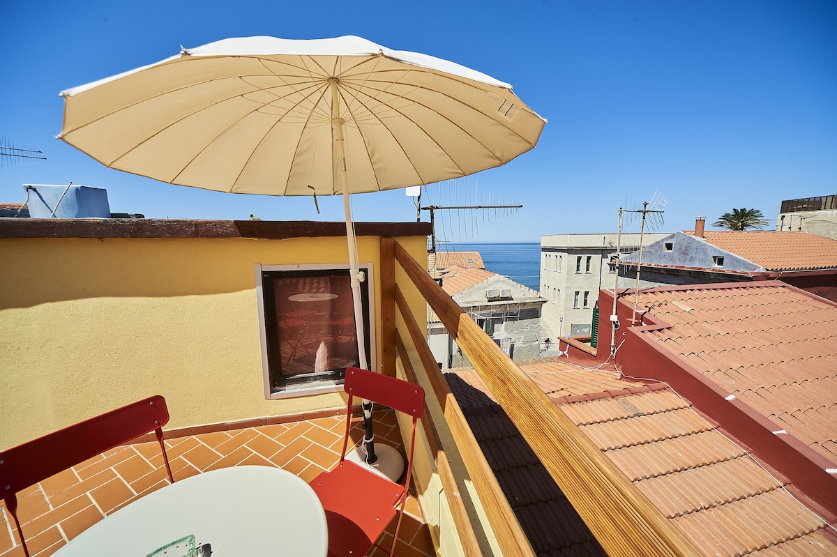 A sunlit terrace features a round table with two red chairs, accompanied by a large umbrella for shade. A view of the rooftops and the sea is visible, presenting a tranquil outdoor space for relaxation.