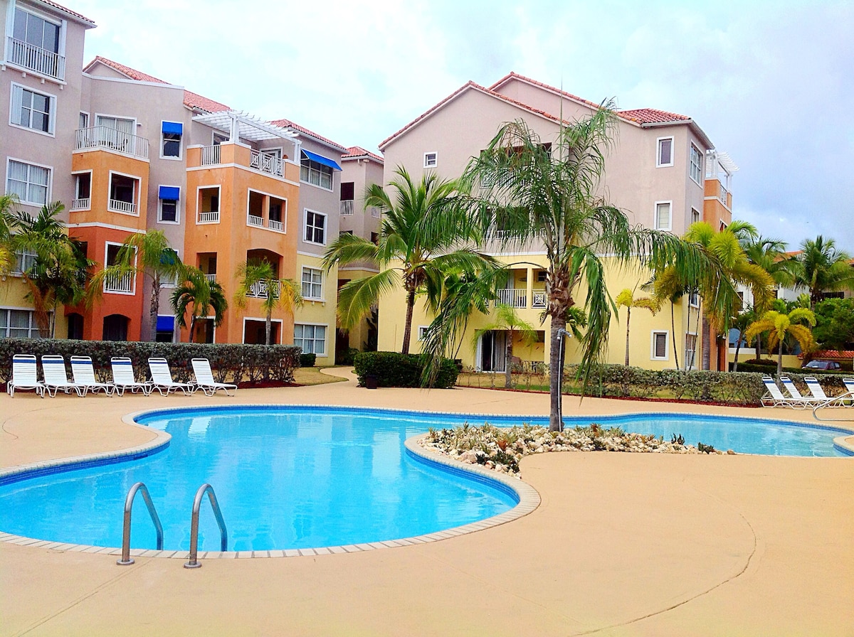 A large swimming pool is surrounded by a tropical landscape featuring palm trees and lounge chairs. The pool's clear water reflects the soft clouds above, while colorful buildings in the background provide a lively atmosphere. The outdoor area invites relaxation and leisure.