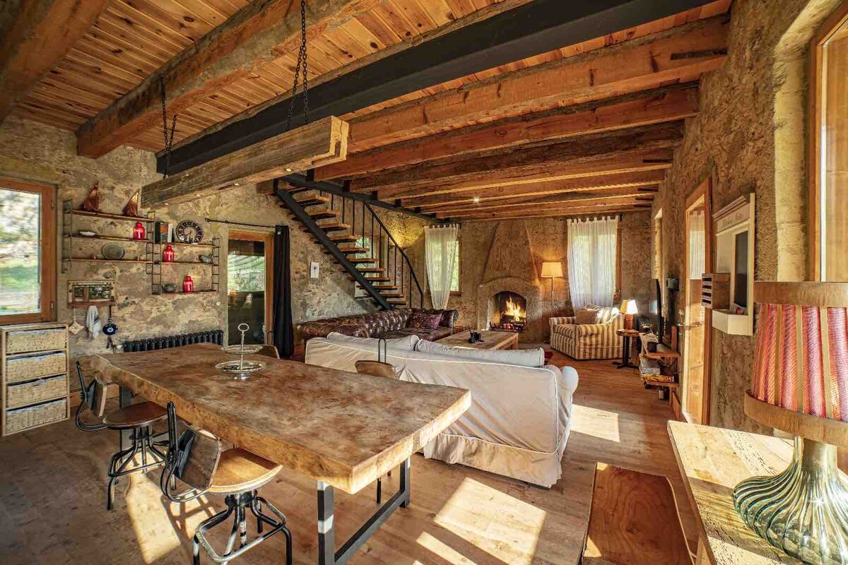 An open living area features exposed wooden beams and a stone wall. A large dining table with metal chairs is positioned near a cozy fireplace, surrounded by comfortable seating. Soft light filters through large windows, illuminating the space. A staircase leads to an upper level.