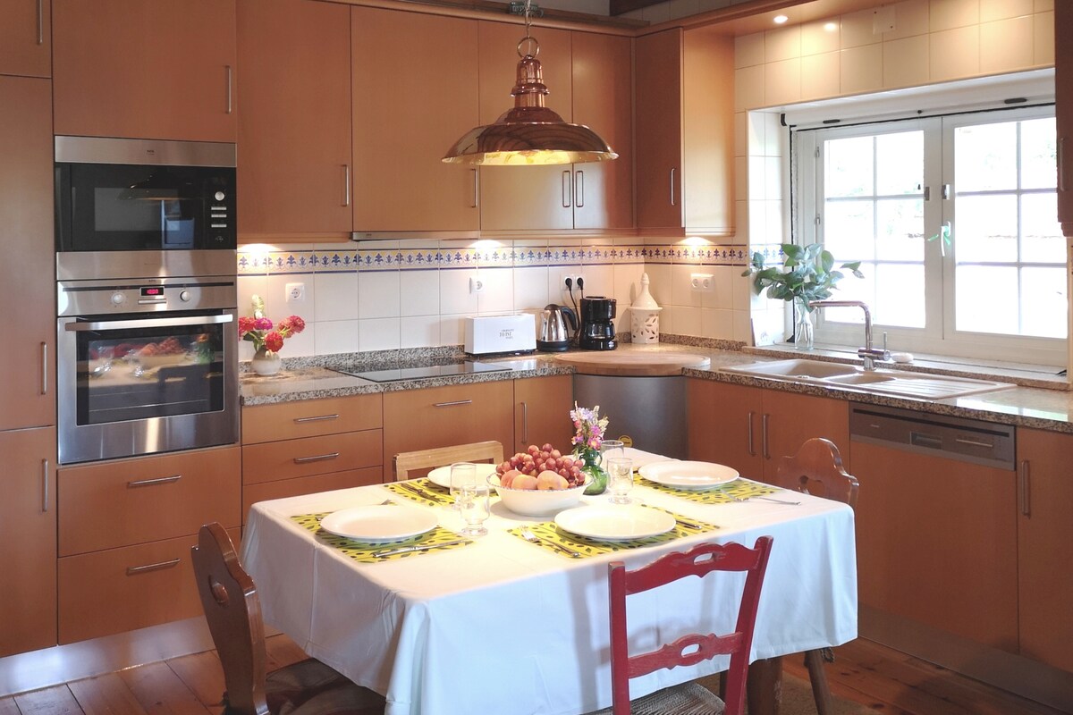 A modern kitchen is presented with wooden cabinetry and sleek appliances. A polished countertop is adorned with a small vase of flowers and a bowl of fruit. A neatly set dining table is centered in the space, featuring white plates and vibrant green placemats.