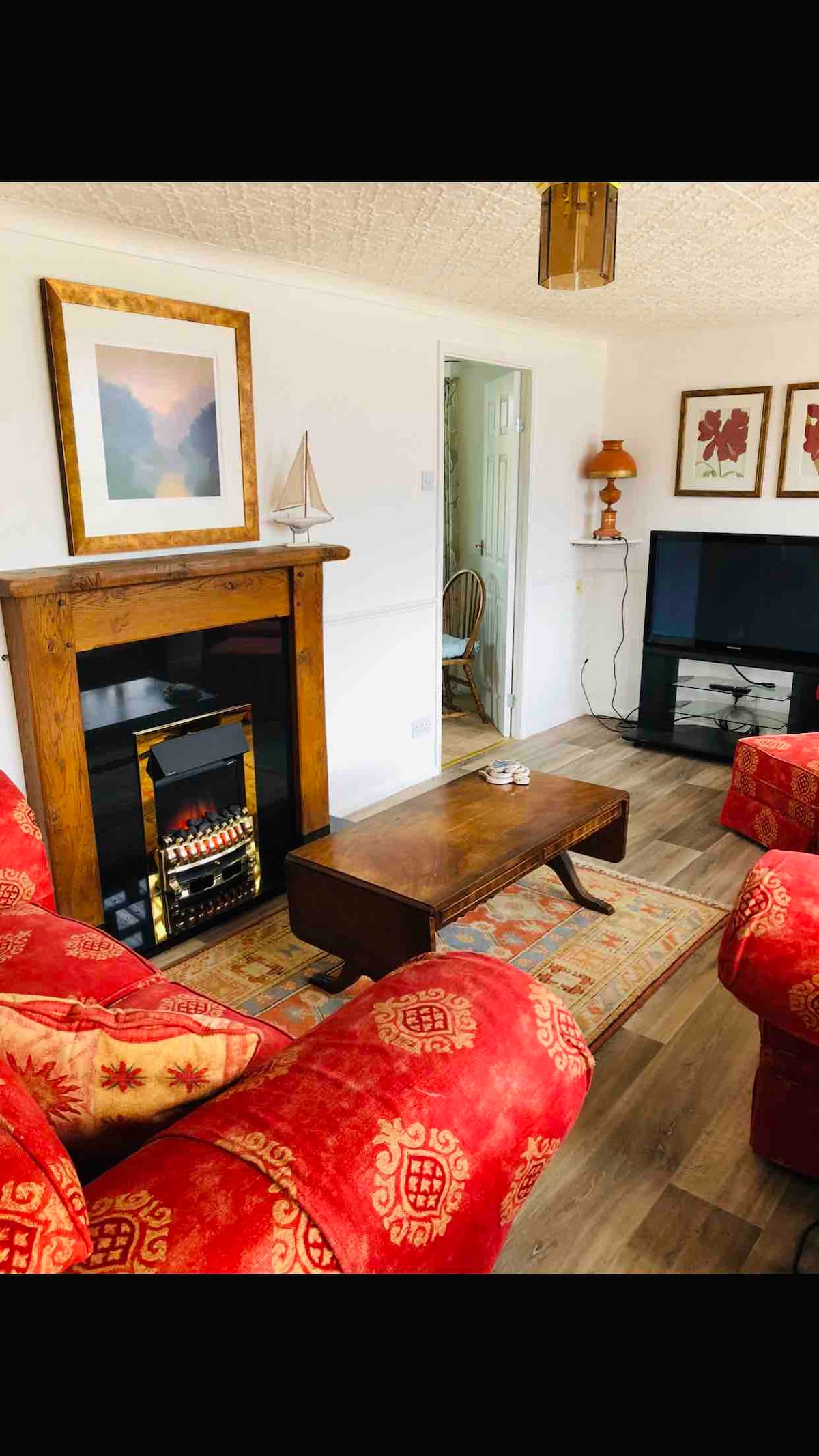 The cozy lounge features a warm wooden fireplace with a decorative sailboat and a vintage lamp. A low wooden coffee table sits on a patterned rug, surrounded by two plush red chairs. A TV and console are positioned against the wall, completing the inviting space.