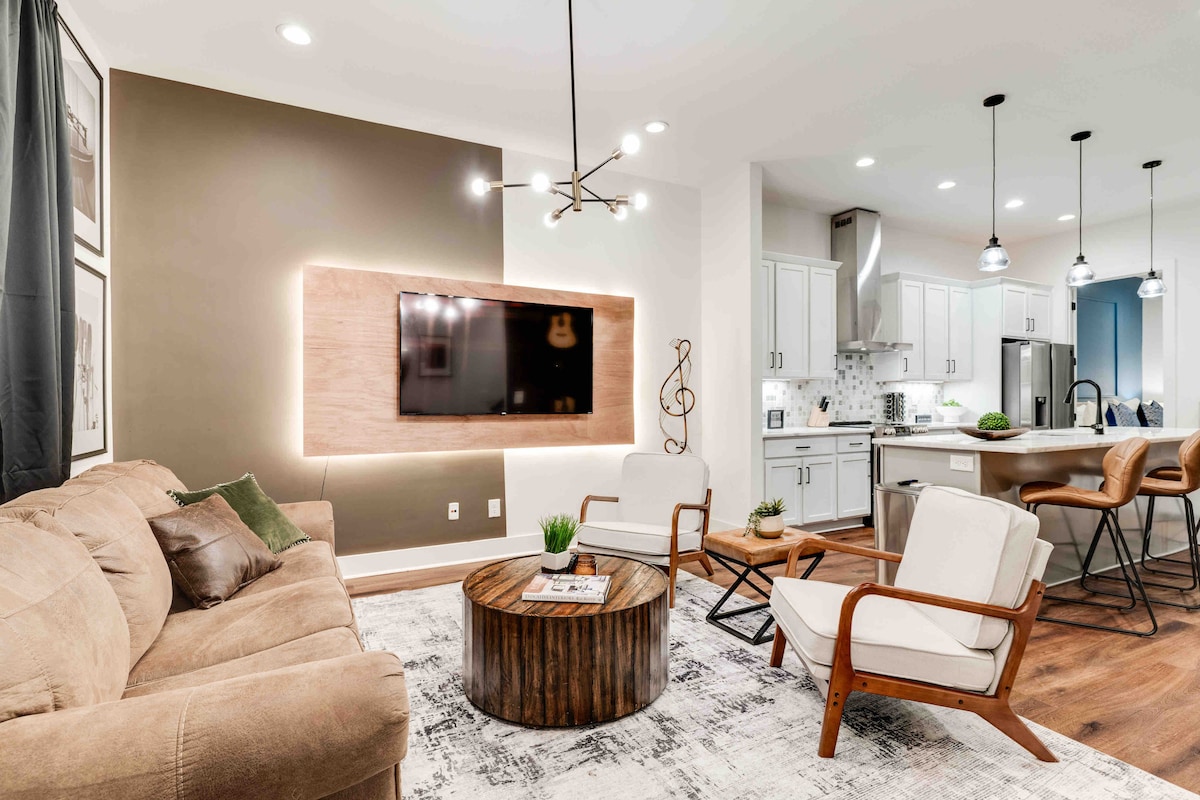 The living area features a soft sofa and two lounge chairs arranged around a rustic wooden coffee table. A mounted television is set against a modern backdrop, while the open kitchen is visible in the background, showcasing sleek cabinetry and stainless-steel appliances.