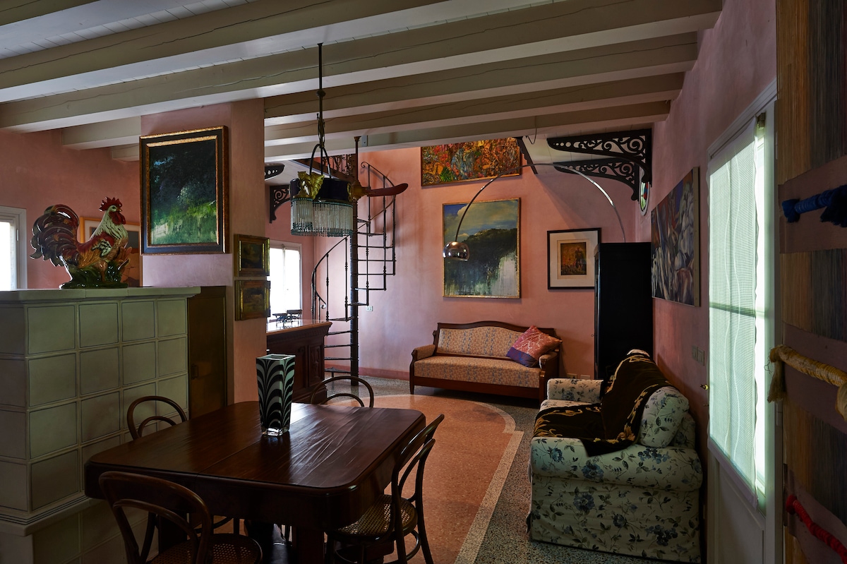 An inviting common area showcases a blend of artistic decor with various paintings on the walls. A wooden dining table is paired with woven chairs, while a comfortable sofa offers seating. Natural light enters from multiple windows, enhancing the spacious atmosphere.