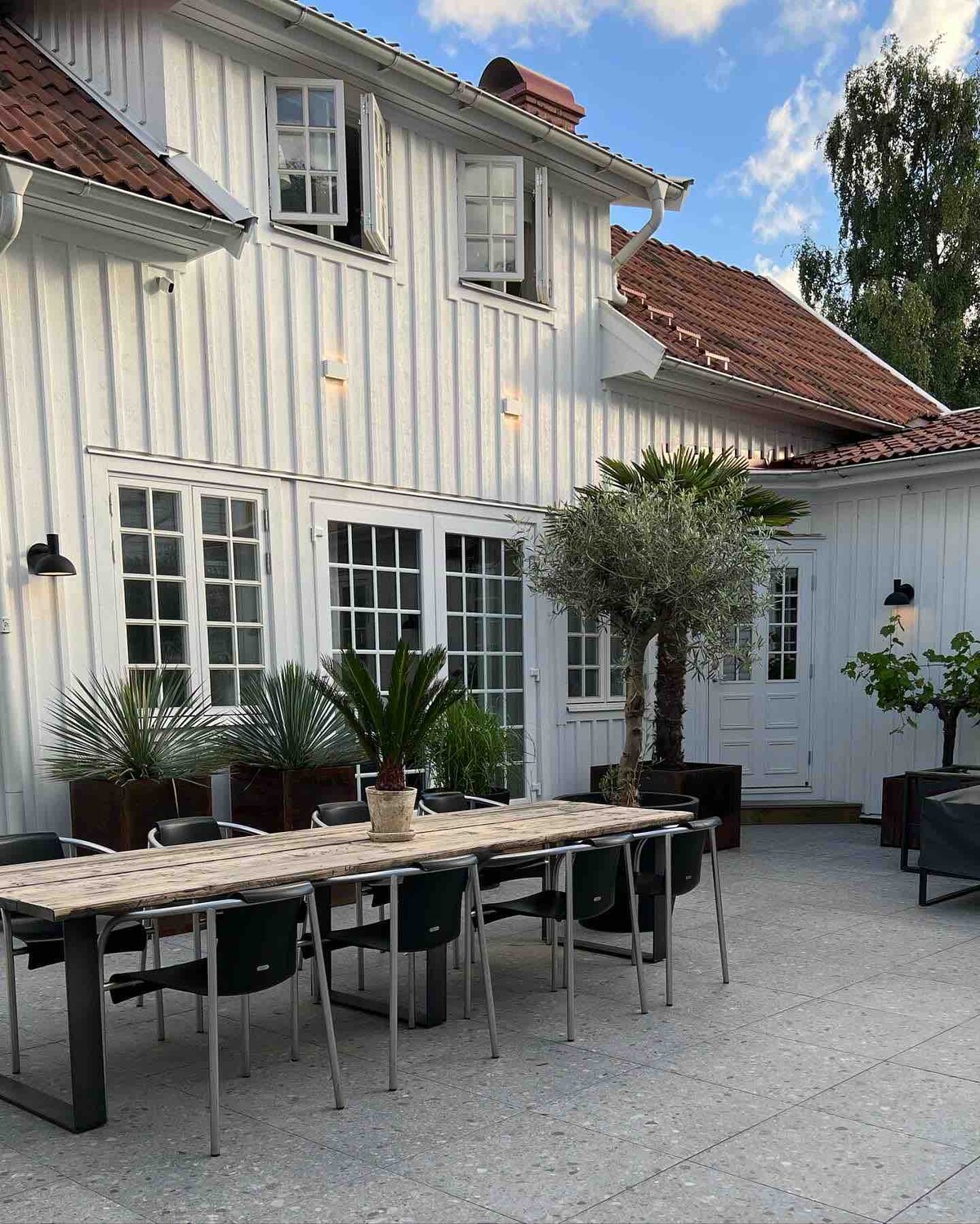 A spacious outdoor patio features a long wooden dining table surrounded by metal chairs. Lush plants and trees add greenery to the area. The white wooden house presents multiple windows and an inviting entrance in the background, creating a serene outdoor setting.