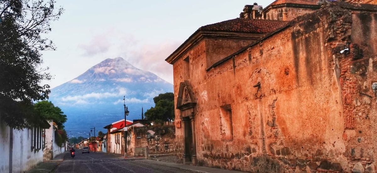 The image displays a historic street in the colonial center of Antigua Guatemala, lined with weathered stone walls and traditional architecture. In the background, a prominent volcano rises under a softly lit sky, offering a striking natural backdrop to the scene.