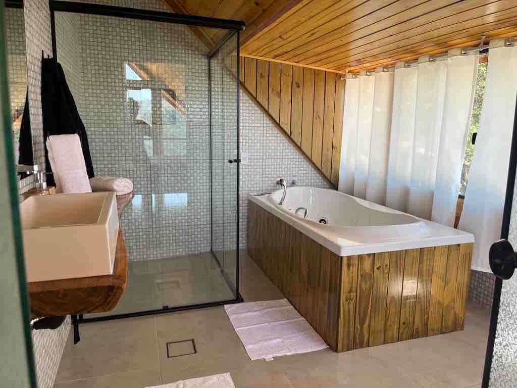A well-lit bathroom features a spacious jacuzzi tub with a wooden surround, complemented by a modern shower with glass enclosure. Subtle mosaic tiles create texture on the walls, while natural light filters through sheer curtains.