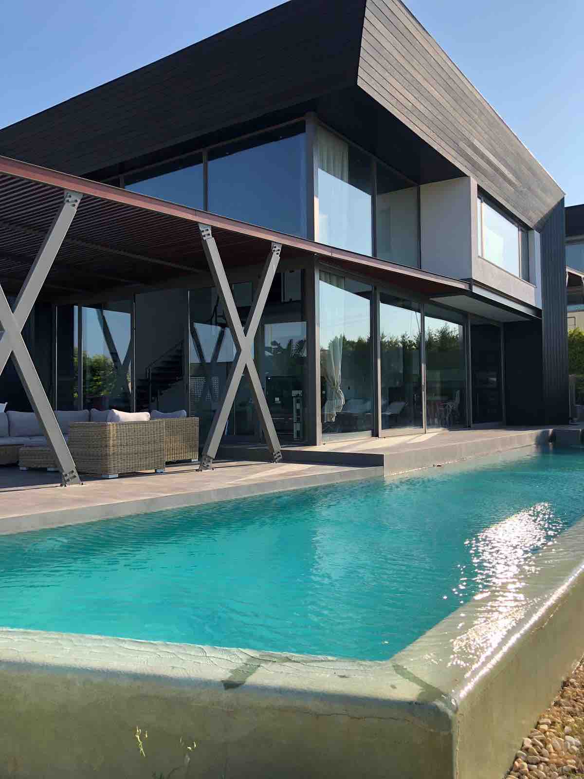 A modern villa exterior features large glass windows reflecting natural light. A private pool with clear blue water is positioned in the foreground, complemented by a wooden terrace area with cozy seating. The architectural design exhibits clean lines and open spaces.