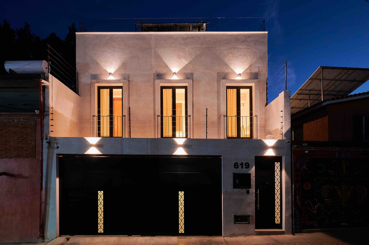 The exterior of the house is illuminated at night, showcasing a modern facade accented by warm lighting. Two balconies with black railings are visible above the entrance. The large garage doors feature decorative designs, adding character to the contemporary structure.