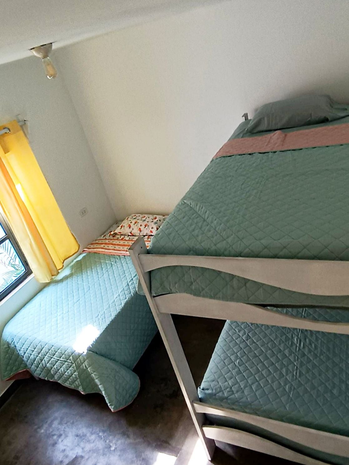 A bright room features a bunk bed and a separate spacious bed. Light yellow curtains frame a window, offering natural light. The bedding is a calming green, and the room displays a minimalist, inviting layout.