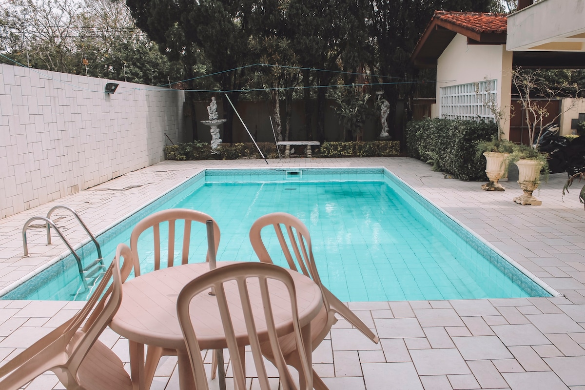 A clear swimming pool is framed by a tiled patio featuring a round table and several light-colored chairs. Lush greenery borders the area, complemented by decorative stone sculptures. Sunlight reflects off the water, creating a tranquil atmosphere in the outdoor space.