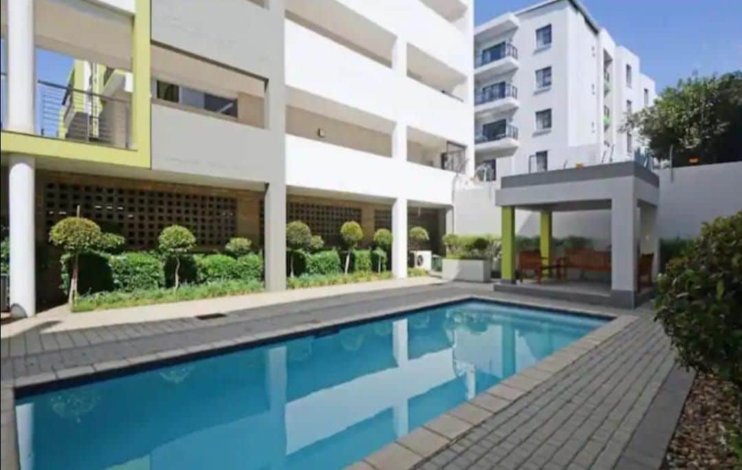 A swimming pool surrounded by carefully maintained greenery is seen. Sunlight reflects off the water's surface, highlighting the clear blue color. Adjacent to the pool, a shaded seating area provides space for relaxation amid the tranquil environment.