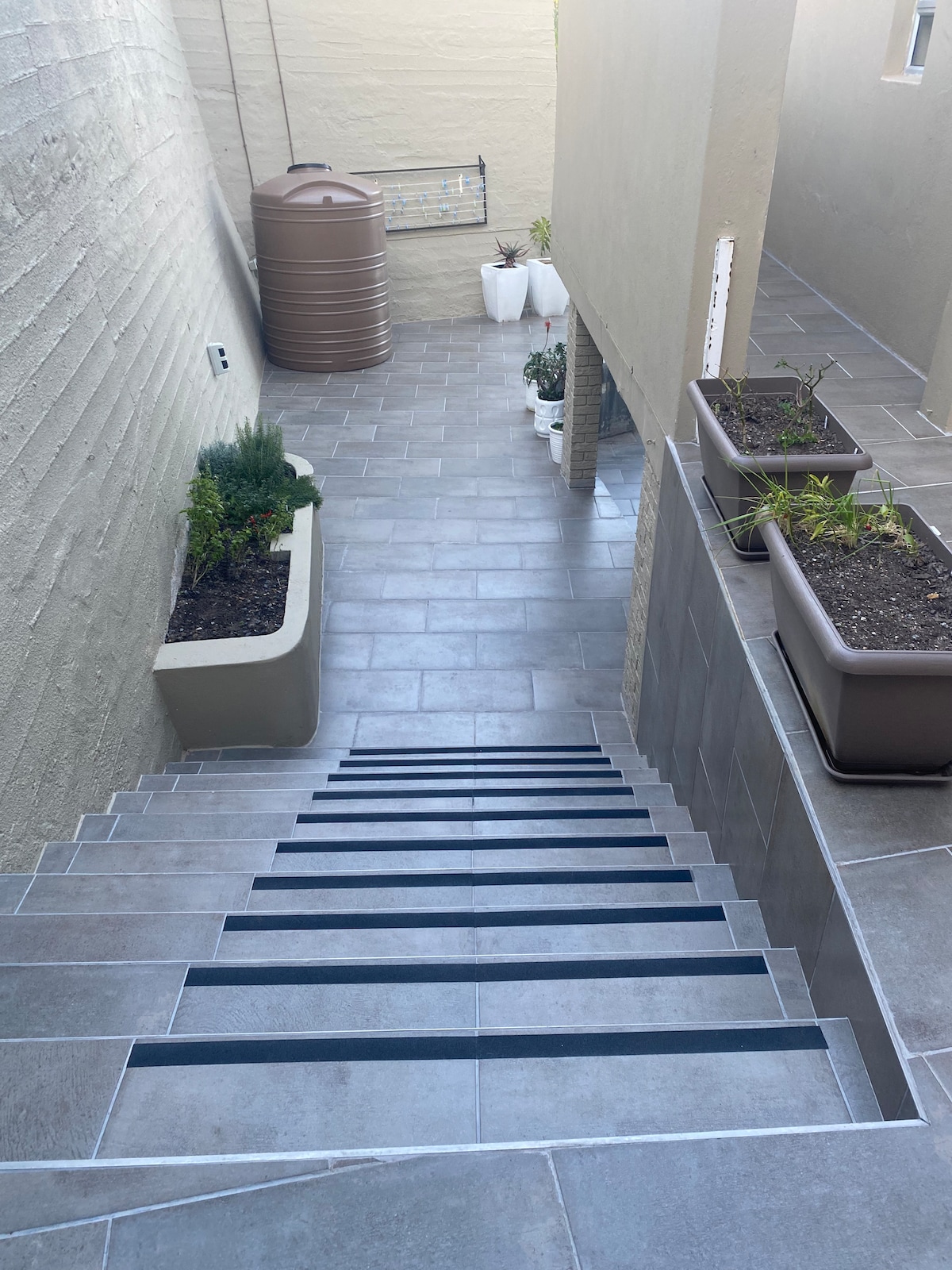 A set of stairs leads down to a tiled pathway surrounded by potted plants. A rainwater tank is visible in the background, alongside gardening supplies. The neutral-toned walls and flooring create a cohesive outdoor space with an accessible and well-maintained appearance.