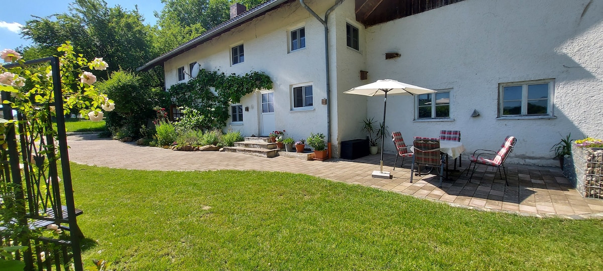 The exterior of the property showcases a well-maintained garden area, featuring a stone patio with a table and chairs shaded by an umbrella. Climbing plants adorn the side of the building, while a grassy lawn extends towards the inviting entrance.