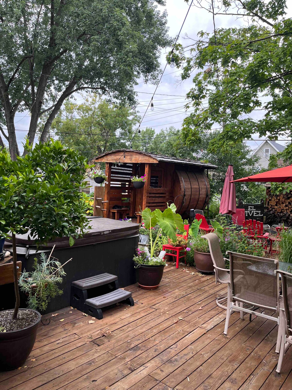 A spacious wooden deck is surrounded by various potted plants and outdoor furniture. A barrel-shaped sauna is visible, alongside a hot tub. Brightly colored umbrellas provide shade over seating arrangements, and lush greenery enhances the outdoor ambiance. A cozy atmosphere is created for relaxation.
