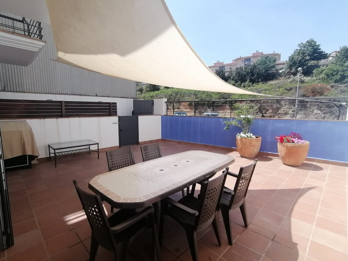 A spacious terrace features a large table surrounded by several chairs, ideal for outdoor dining. The area is shaded by a canopy, with potted plants adding a touch of greenery. A vibrant blue wall provides a colorful backdrop, enhancing the outdoor experience.