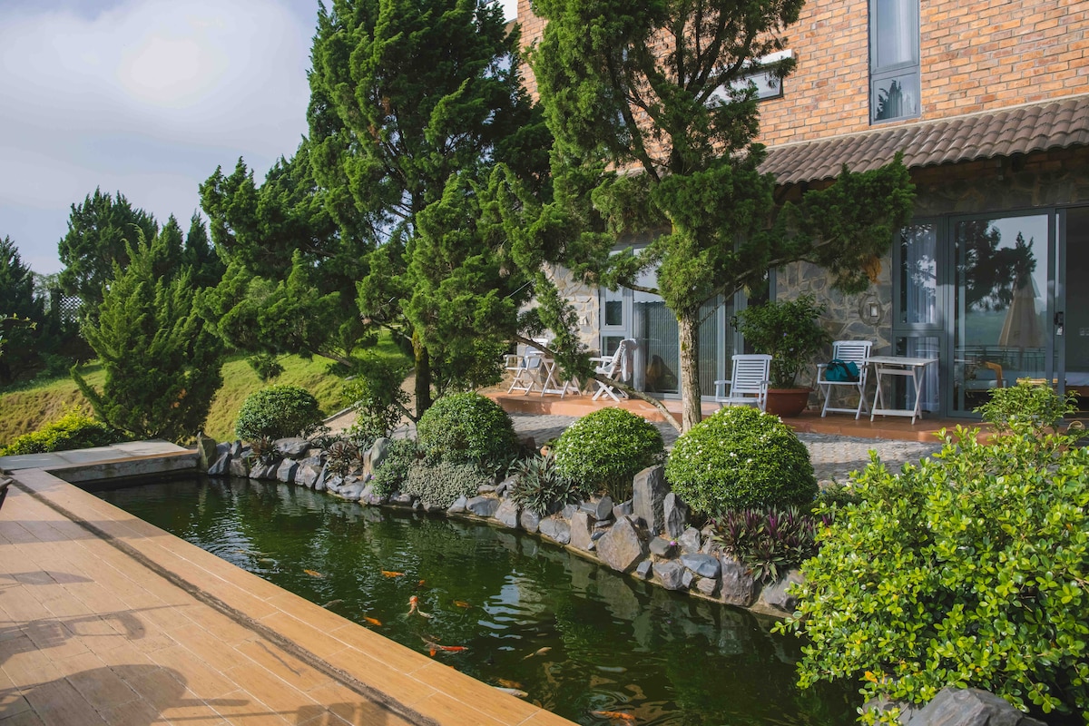A tranquil outdoor area features a pond surrounded by lush greenery and carefully arranged plants. The wooden deck extends towards the water, with white chairs and tables positioned to offer serene views of the garden and hillside.