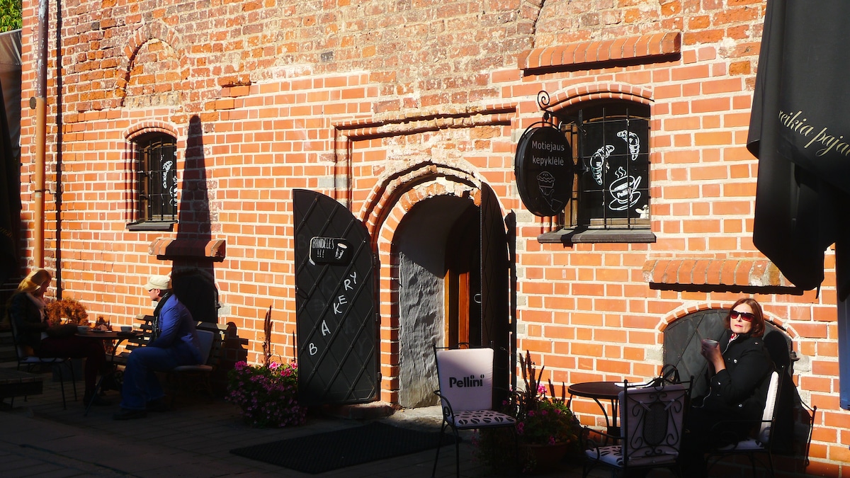 The image captures a charming brick facade, characteristic of Kaunas's old town. An inviting entrance with a dark wooden door leads inside. Tables with chairs are placed outside, accompanied by lively planters. Sunlight casts soft shadows, enhancing the historic feel of the setting.