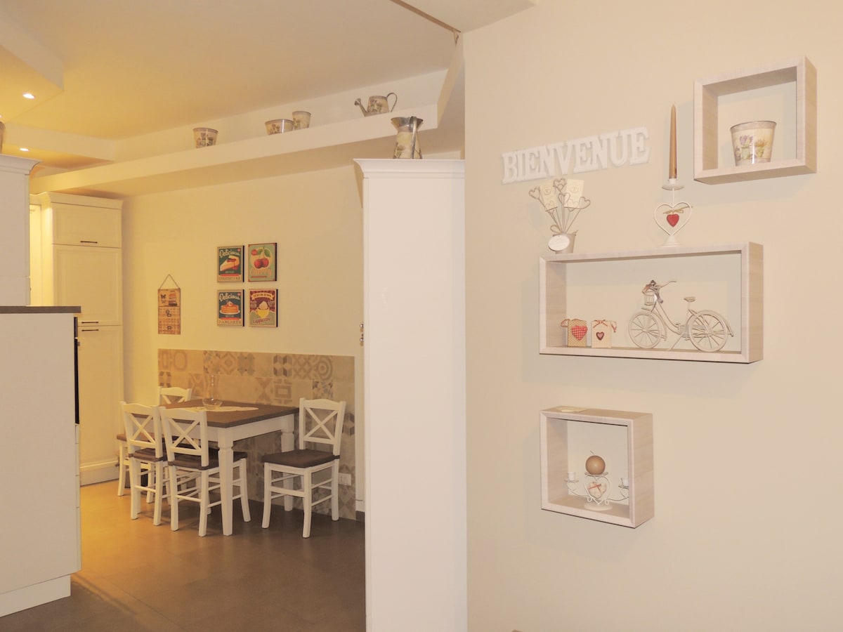 A cozy dining area is visible, featuring a wooden table surrounded by white chairs. Decorative shelving displays charming items, including small figurines and art pieces. Neutral walls create a warm backdrop, complemented by soft overhead lighting that enhances the welcoming atmosphere of the space.