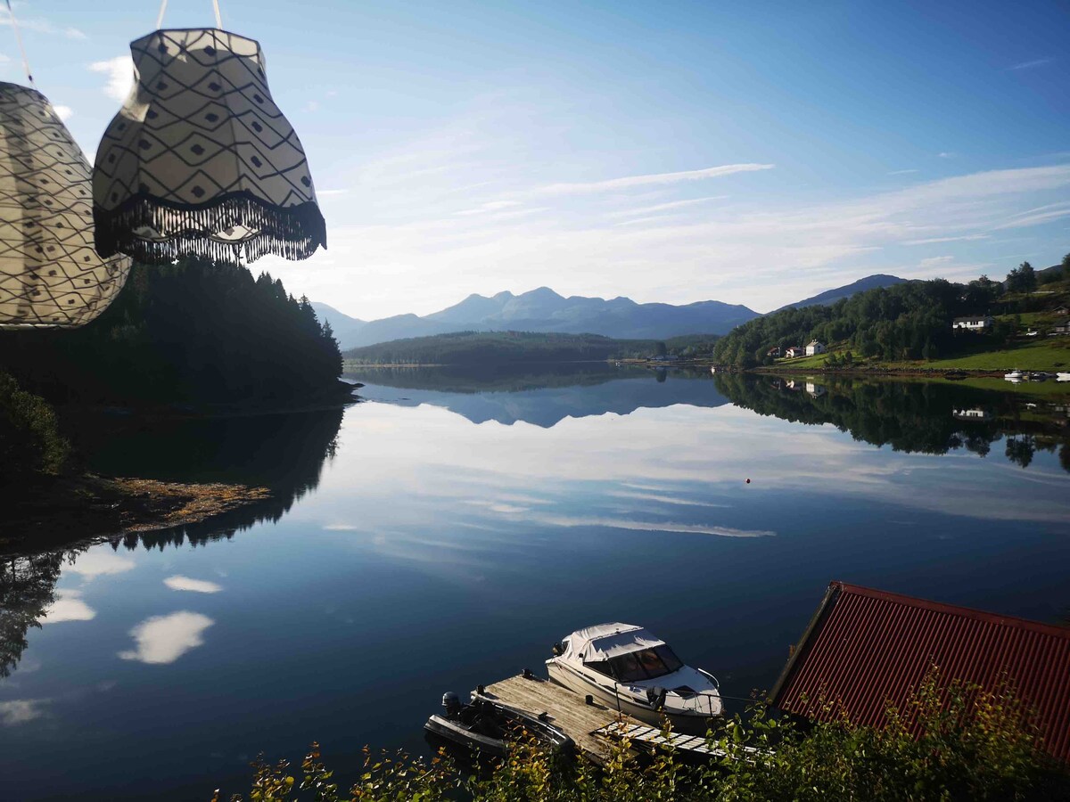 A serene view of a calm fjord reflects the surrounding mountains and clear blue sky. A covered boat rests on a dock with a red-roofed structure in the foreground. Hanging light fixtures add a decorative touch to the scene.