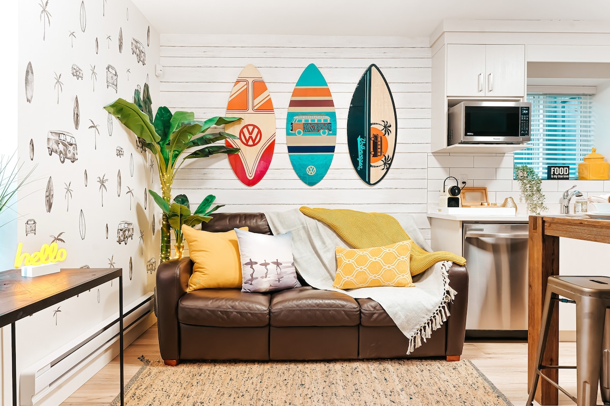 A cozy living area features a leather couch adorned with colorful throw pillows and a soft blanket. Surfboards are mounted on the wall, complemented by tropical plants. A small table and an open kitchen space with modern appliances add to the inviting ambiance.