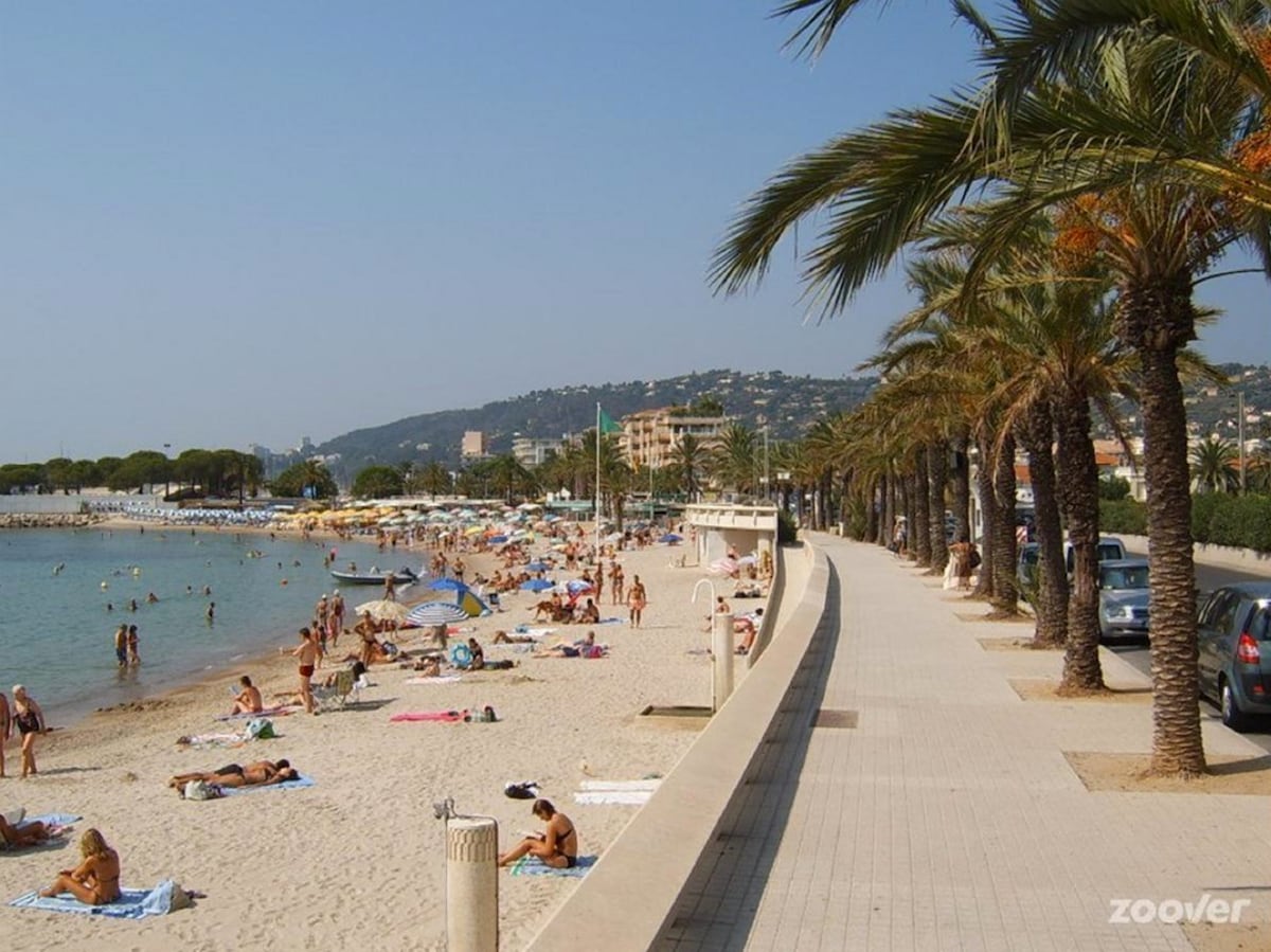 A sandy beach is lined with palm trees, where sunbathers relax on colorful towels. The promenade runs alongside the coast, featuring a smooth walkway and small boats visible in the shallow water. Rolling hills and buildings can be seen in the background.