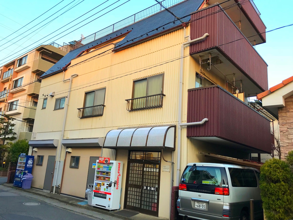 An exterior view of a three-story building is presented, featuring a combination of beige and maroon siding. Large windows are visible on each floor, while a covered entrance is situated in the center. An adjacent vehicle and vending machine contribute to the urban setting.